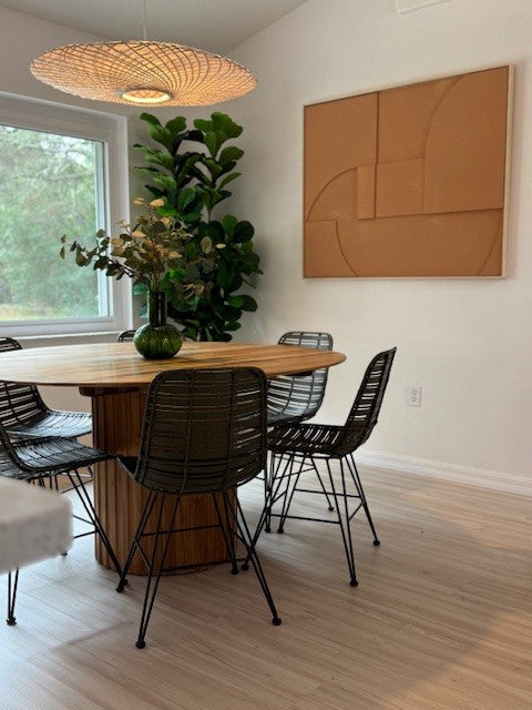 A modern dining room scene featuring a round wooden dining table surrounded by black wire chairs, with a large woven pendant light overhead and a cork wall art piece on the white wall.