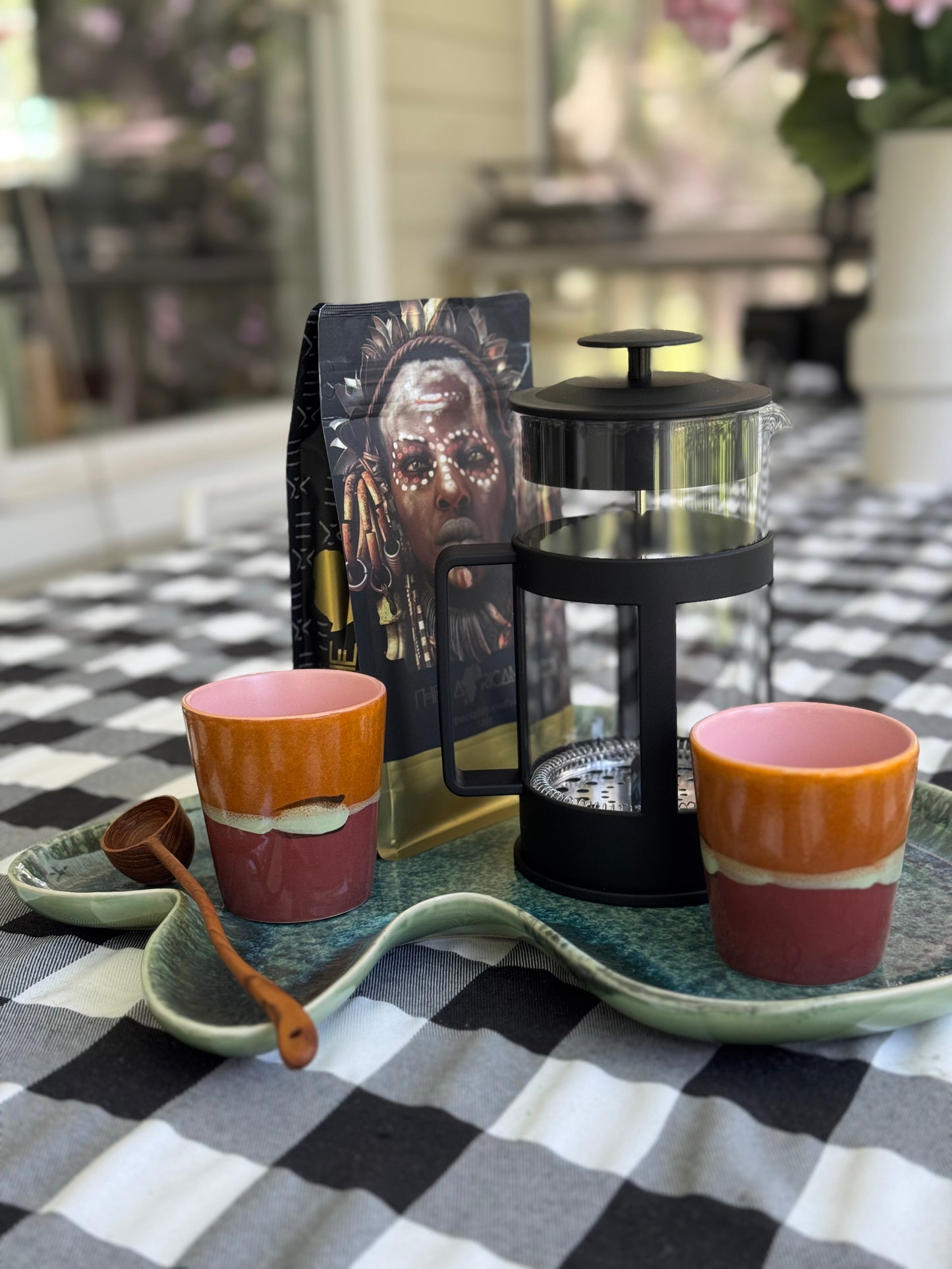 French press coffee maker with two colorful mugs on a checkered tablecloth.