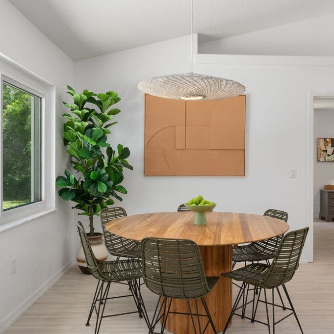 A modern dining room featuring a round wooden dining table surrounded by six woven dark green chairs, under a textured pendant light. The room has white walls, a large abstract orange-brown wall art piece, a tall potted green plant, and light wood flooring.